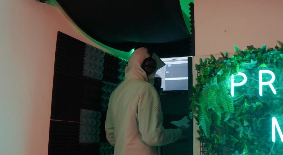 Young person recording in the booth — neon lighting, acoustic panels, Logic Pro on screen